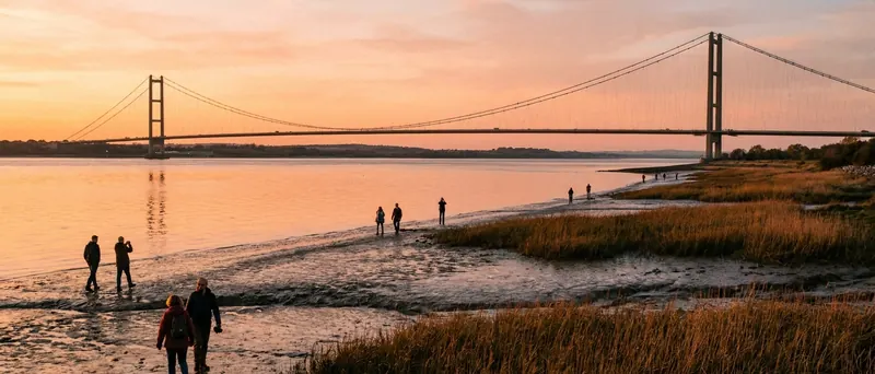 A wide landscape photograph of people walking along muddy foreshore and reeds with a large suspension bridge spanning a wide estuary at sunset.